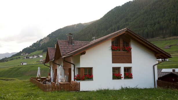 A white house with a sloped roof, wooden balcony, and red flowers in the windows. - Apartment "Rieglhof Maisel" with Mountain View, Garden & Wi-Fi (Graun im Vinschgau)