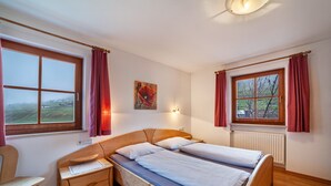 A bedroom with a wooden bed, a nightstand, a chair, two windows with curtains, and a painting on the wall. - Apartment "Rieglhof Maisel" with Mountain View, Garden & Wi-Fi (Graun im Vinschgau)