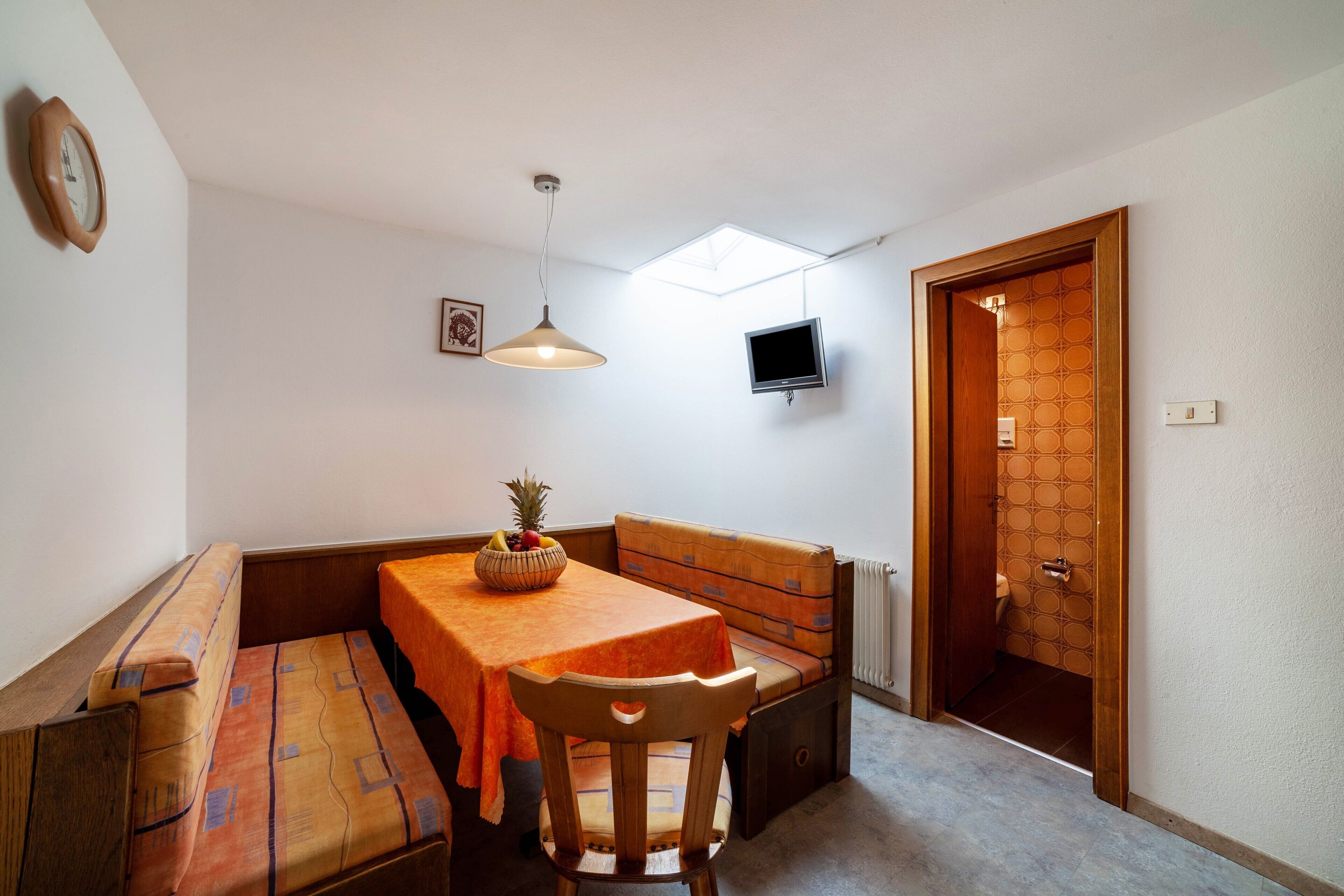 Una habitación compacta con una mesa de comedor, asientos tipo banco, un televisor y una puerta que da a un baño.