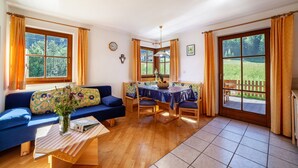 A room with a blue sofa, a wooden dining table with chairs, a vase with flowers, and large windows with curtains. - Apartment 'Rieglhof Geiernest' with Mountain View, Garden & Wi-Fi (Graun im Vinschgau)