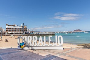 A beachside sign with 'CORRALEJO' prominently displayed, a marina with boats, and a building with multiple floors in the background.