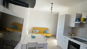 A compact kitchen with white cabinetry, a dining table with chairs, and a yellow sofa with patterned cushions. - STILL HOUSE (San Nicola Arcella)