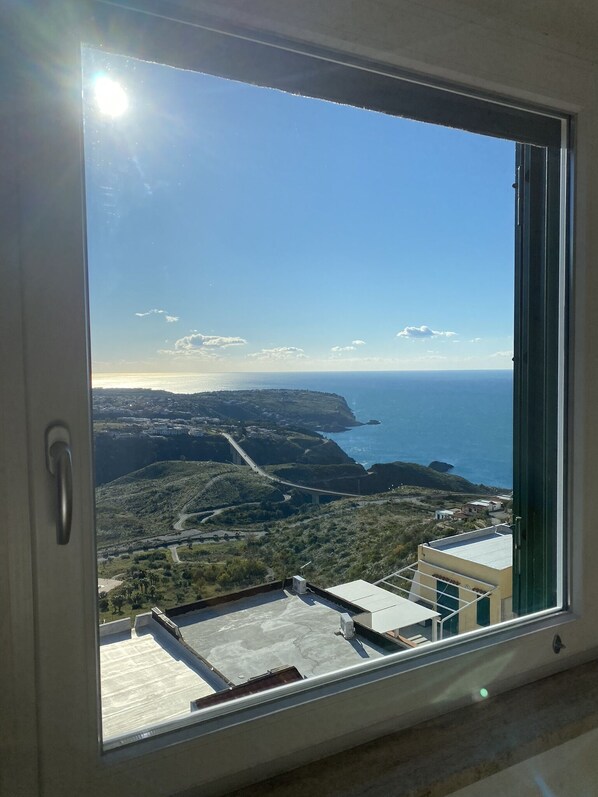 A room with a large window overlooking a coastal landscape, including a bridge and distant hills. - STILL HOUSE (San Nicola Arcella)