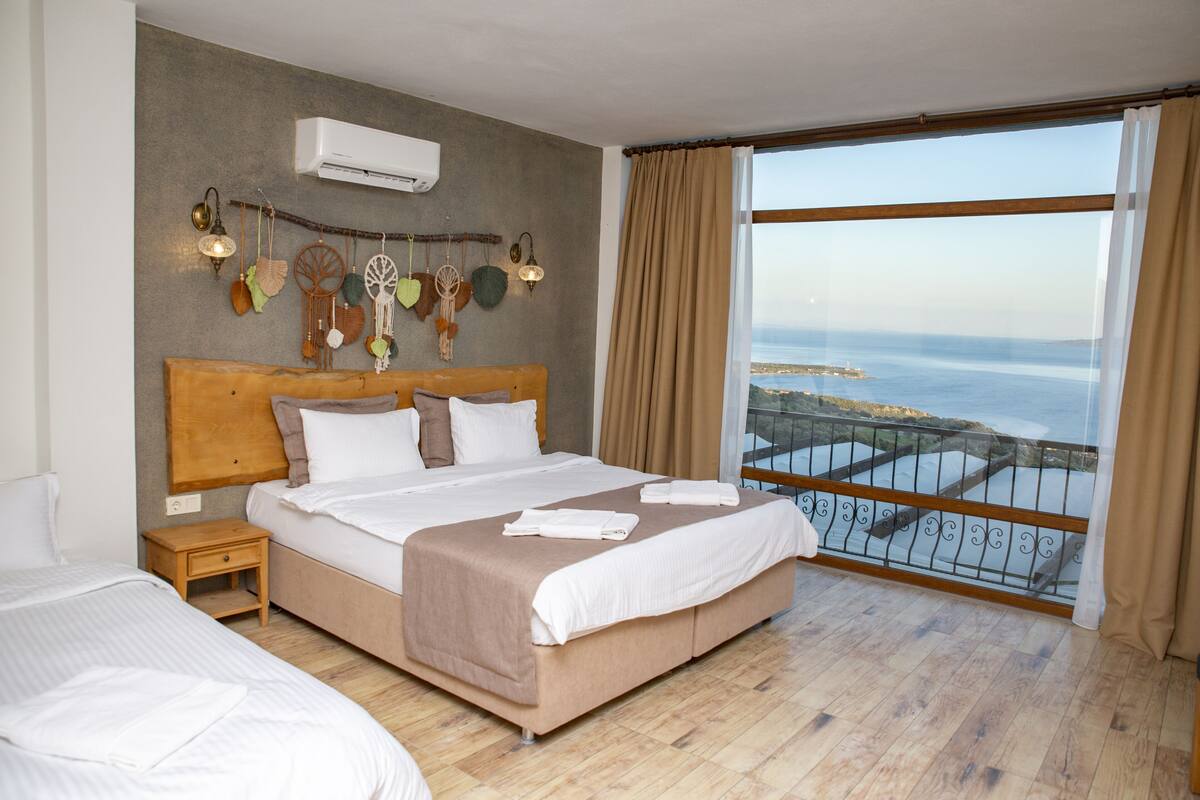A bedroom with a large bed, a nightstand, a wall-mounted air conditioner, and a balcony with a view of the sea.