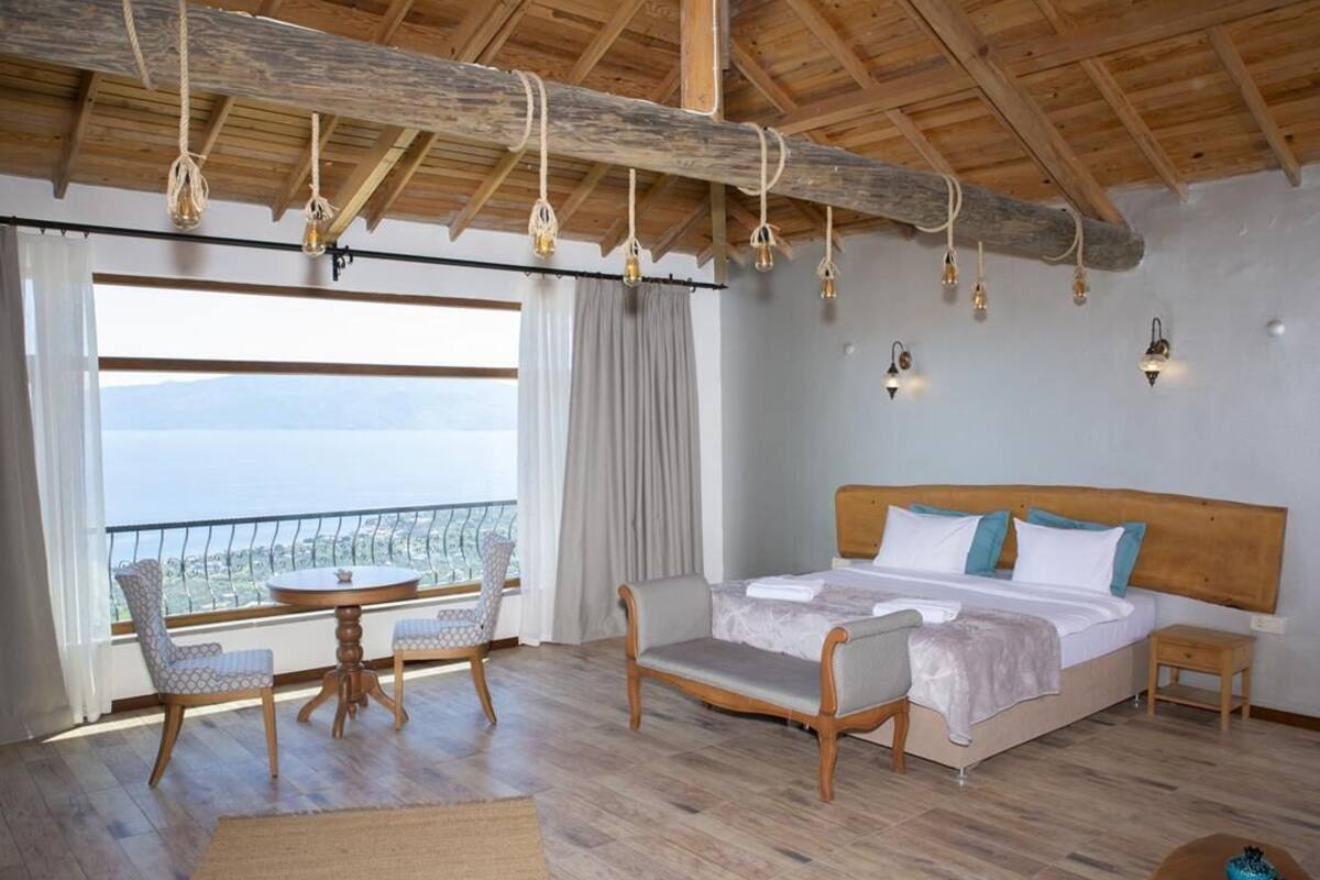 A room with a bed, a small table, two chairs, a view of the sea, and a wooden ceiling.
