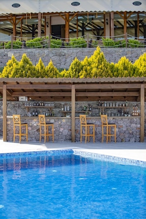 A poolside bar with wooden chairs and a stone wall backdrop, overlooking a swimming pool.