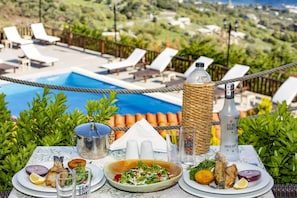 A dining table with plates of food, a bottle of Yeni Raki, and a view of a pool and lounge chairs.