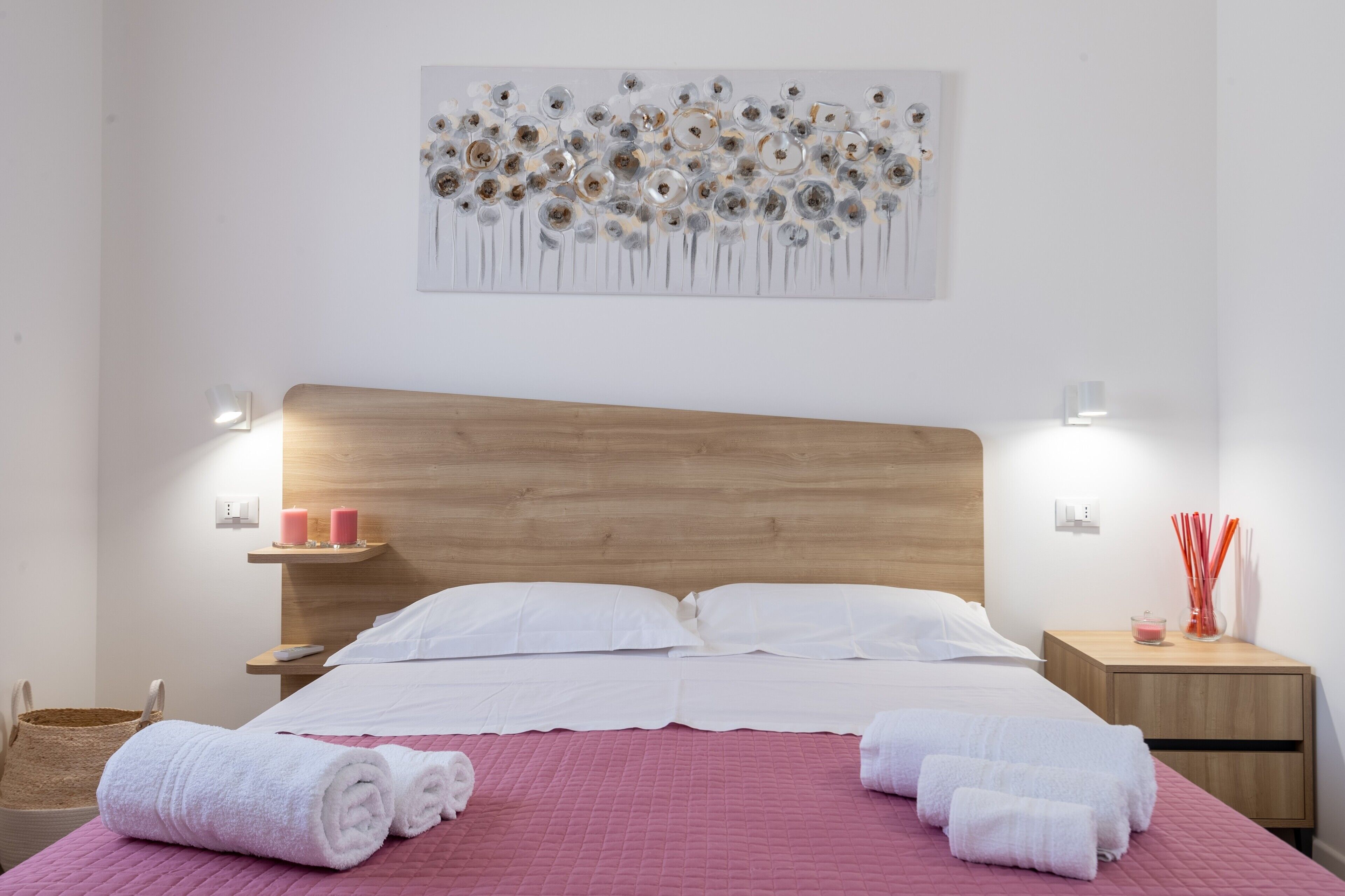 Ein ordentlich bezogenes Bett mit weißen Leinen, einem Holzkopfstück und einer gesteppten rosa Bettdecke. Zwei Nachttische mit Lampen und ein an der Wand hängendes Kunstwerk befinden sich darüber.