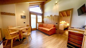 A room with a wooden table and chairs, a sofa, a bed with a nightstand, a TV, and a window with curtains. - Flat Paradisin - Baita Cusini with mountain view, wellness area and Wi-Fi (Livigno)