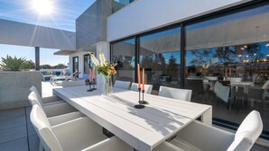 A modern outdoor dining area with a white table and chairs, a glass partition, and a view of the surrounding landscape.