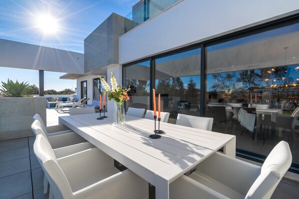A modern outdoor dining area with a white table and chairs, a glass partition, and a view of the surrounding landscape.