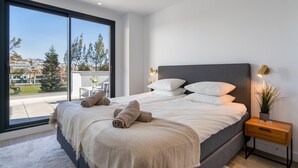 A bedroom with a large window, a bed with white linens, a bedside table with a lamp, and a view of a balcony with outdoor furniture.