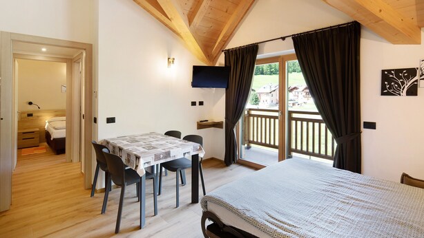 A bedroom with a bed, a table with chairs, a TV, and a balcony with curtains.