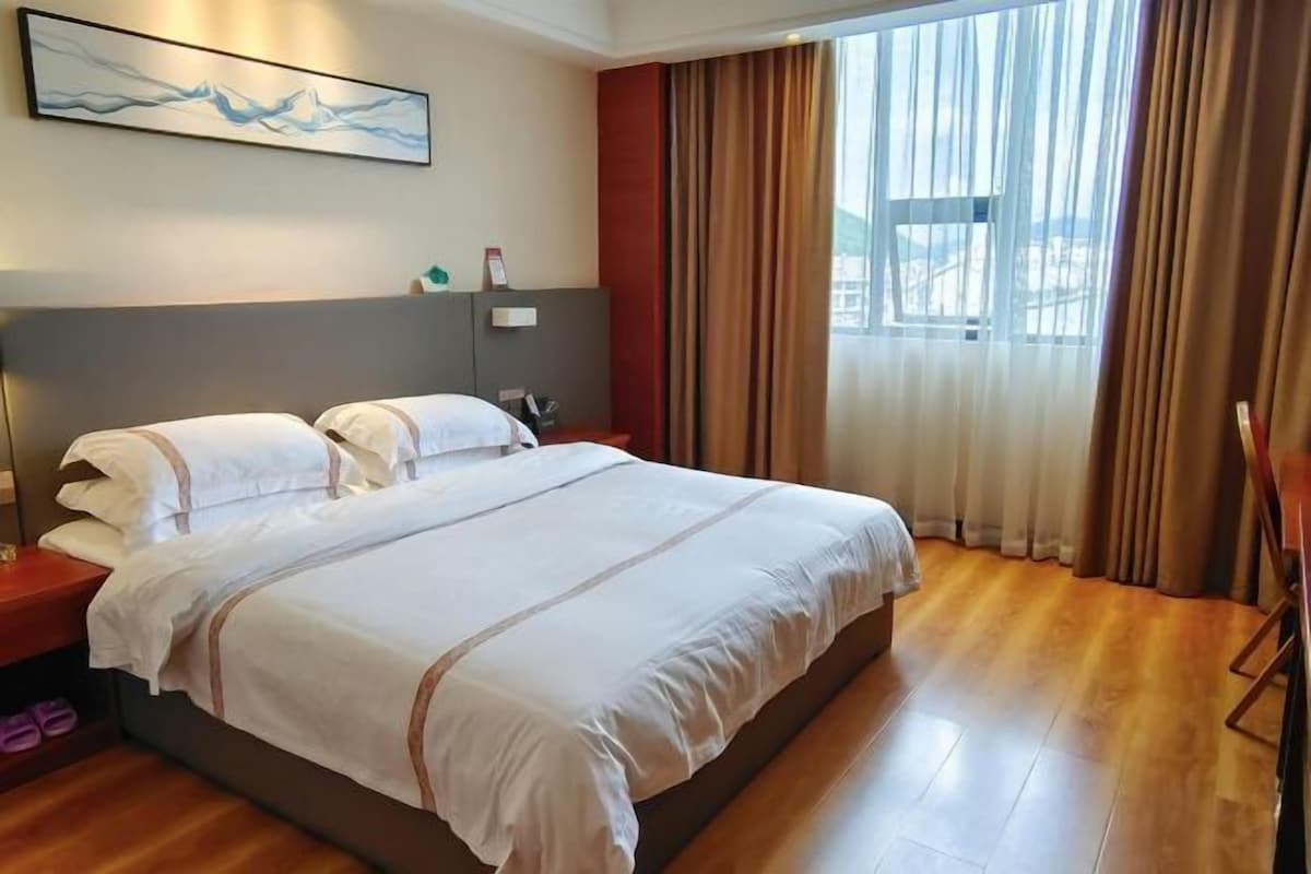 Hotellihuone, jossa on suuri sänky, yöpöytä, lamppu, seinällä oleva taulu ja ikkuna, jossa on verhot.