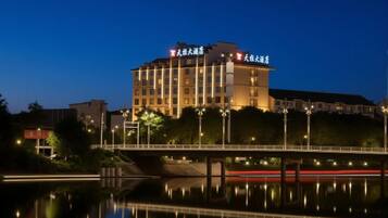 A multi-story hotel with a bridge in the foreground, a body of water reflecting the building, and streetlights along the bridge.