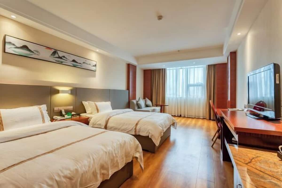 Ein Hotelzimmer mit zwei Betten, einem Schreibtisch mit Fernseher, einem Bild an der Wand und einem Fenster mit Vorhängen.