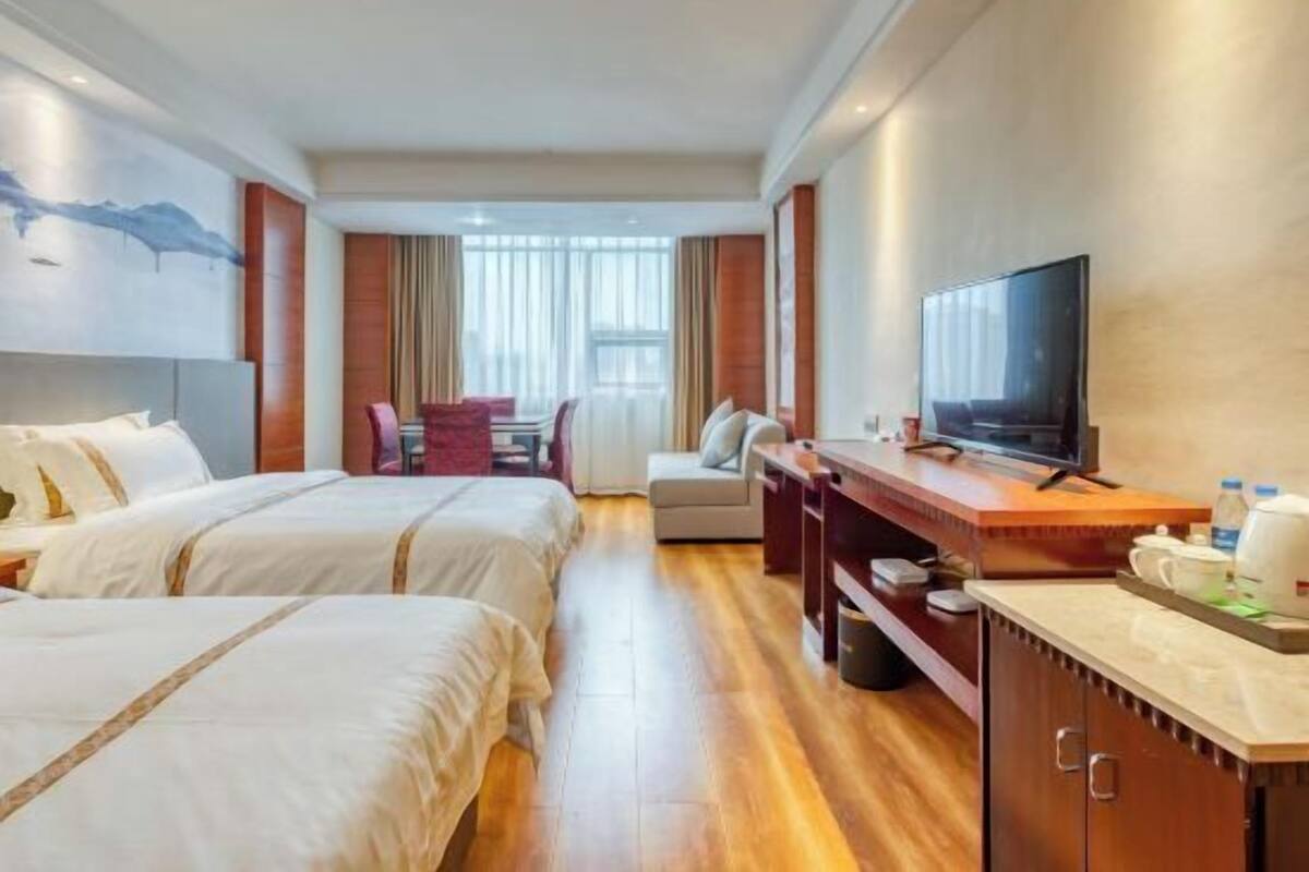 Ein Hotelzimmer mit zwei Betten, einem Schreibtisch mit Fernseher, einem Esstisch mit Stühlen und einem großen Fenster mit Vorhängen.
