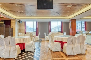 Un salón de banquetes con mesas redondas, sillas blancas y vistas a la ciudad a través de amplios ventanales.