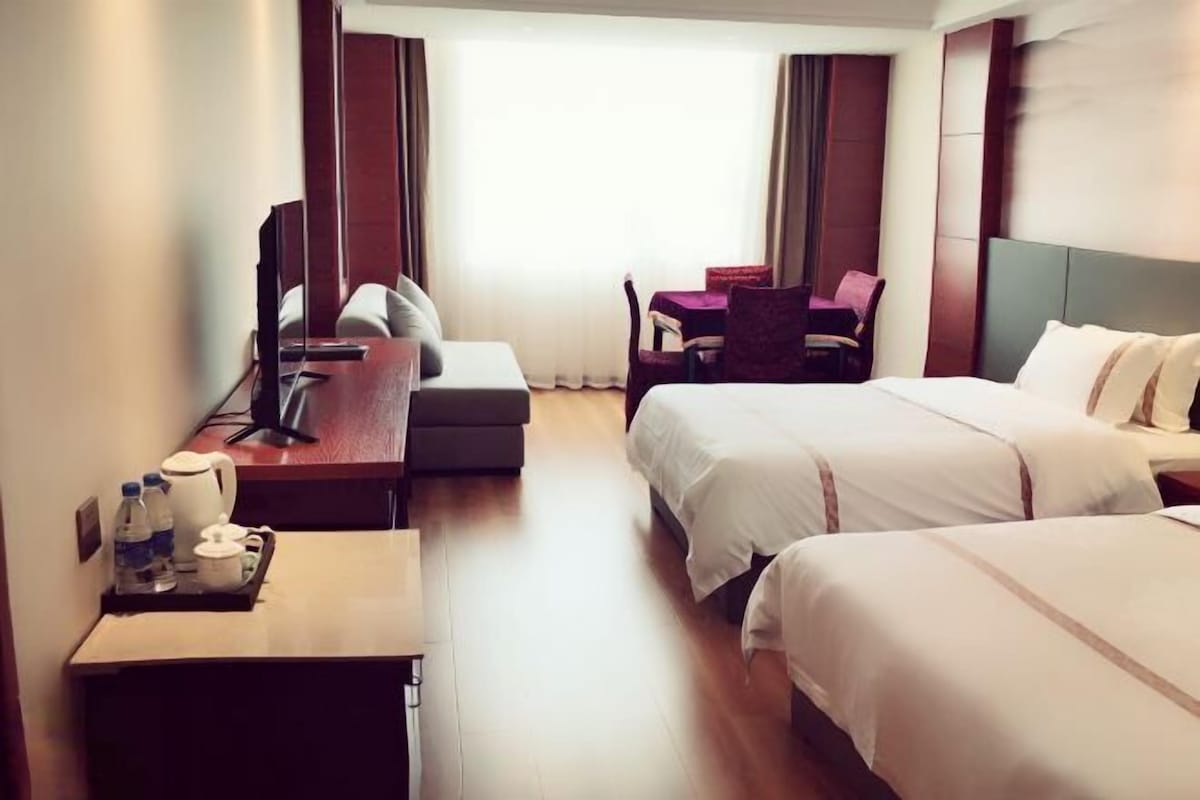 Hotellihuone, jossa on kaksi sänkyä, työpöytä tietokoneella, tuoli ja pieni pöytä, jonka päällä on tarjotin, jossa on erilaisia esineitä.