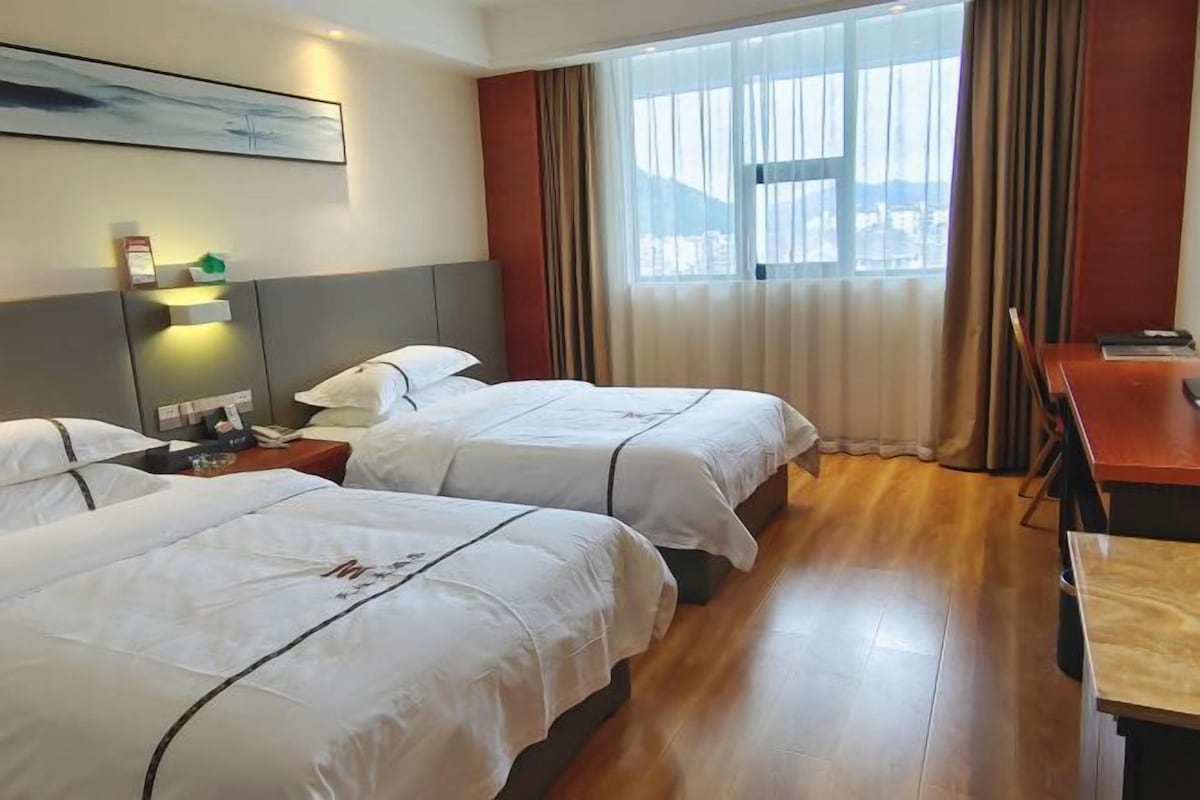 Hotellihuone, jossa on kaksi sänkyä, työpöytä, tuoli, ikkuna, jossa on verhot, taulu seinällä sekä yöpöytä, jossa on lamppu ja puhelin.