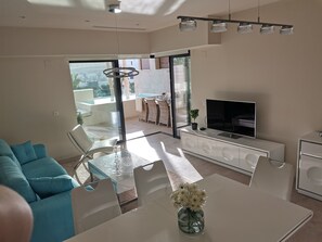 A modern living room with a glass dining table, a sofa, a TV, and a balcony with a pool.