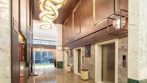 Un moderno vestíbulo de hotel con piso de mármol, una gran lámpara decorativa, ascensores y una puerta de vidrio que da al exterior.