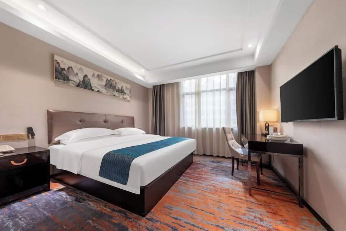 A hotel room with a bed, desk, chair, TV, artwork, and a large window.