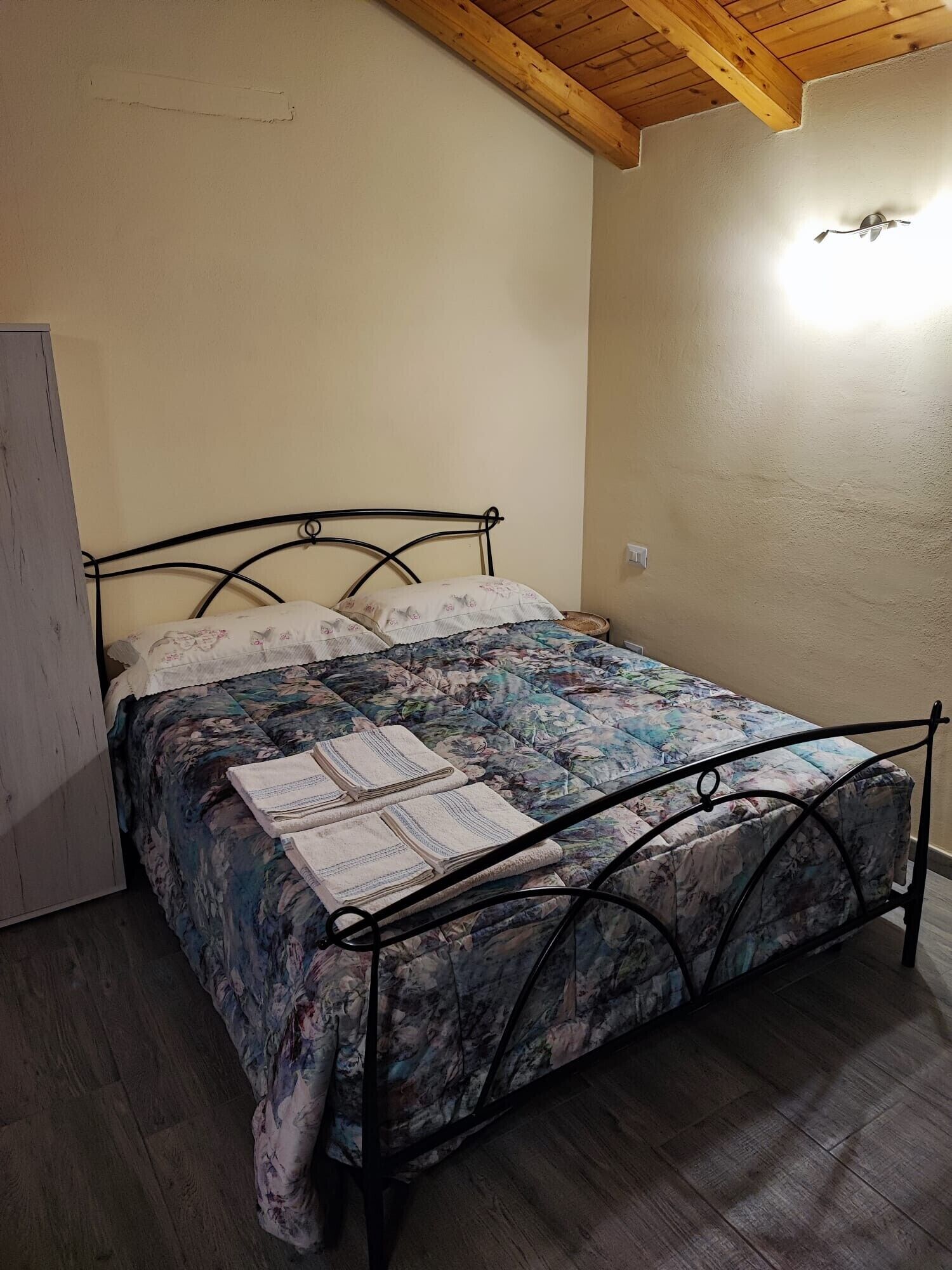 A single bed with a floral bedspread and pillows, a wooden ceiling, and a white wall.