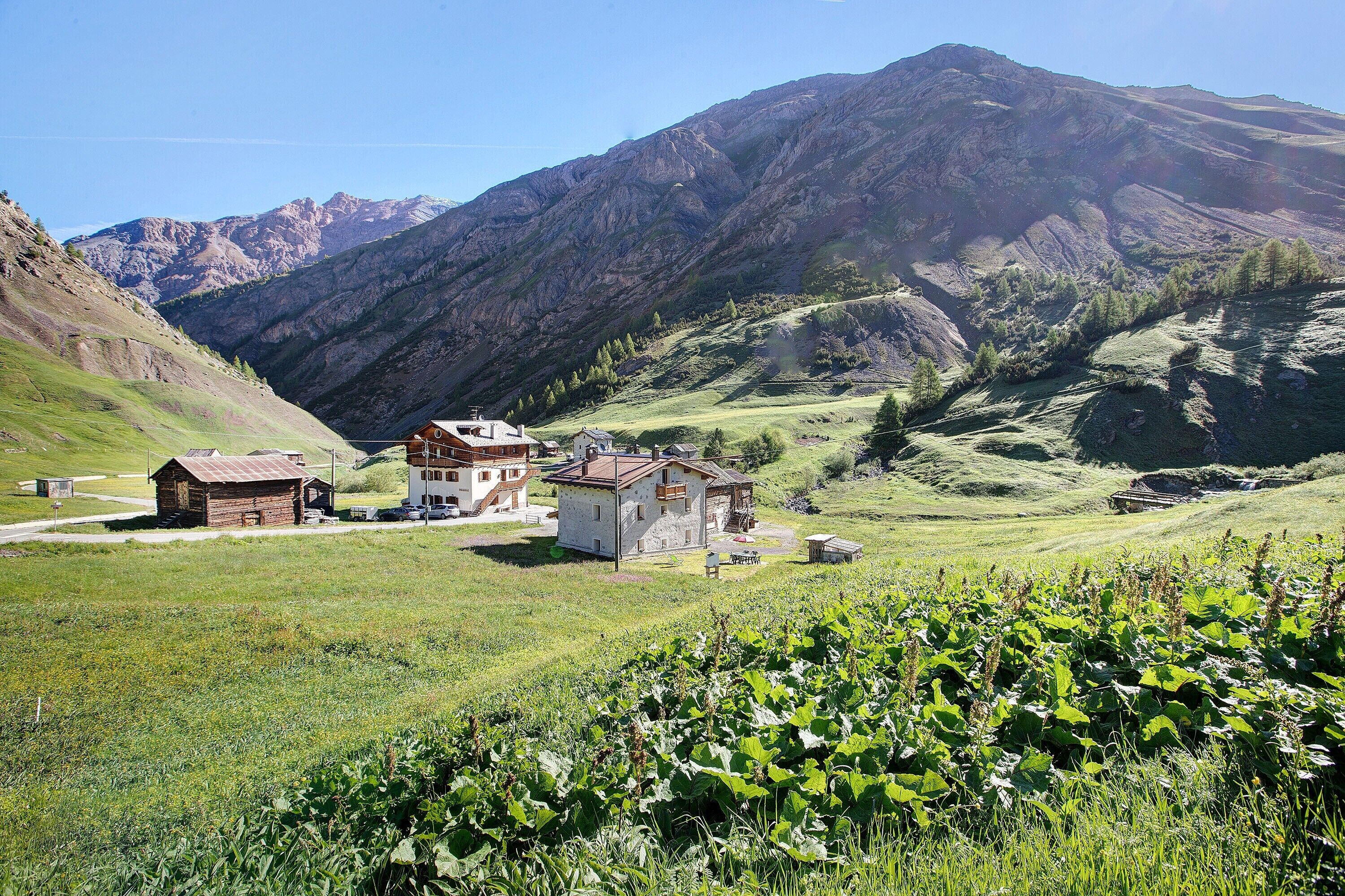 A rural landscape with a mountain backdrop, featuring a small cluster of buildings with a dirt path leading to them.