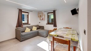 A living room with a sofa, a dining table with chairs, a flat-screen TV mounted on the wall, and a window with curtains.