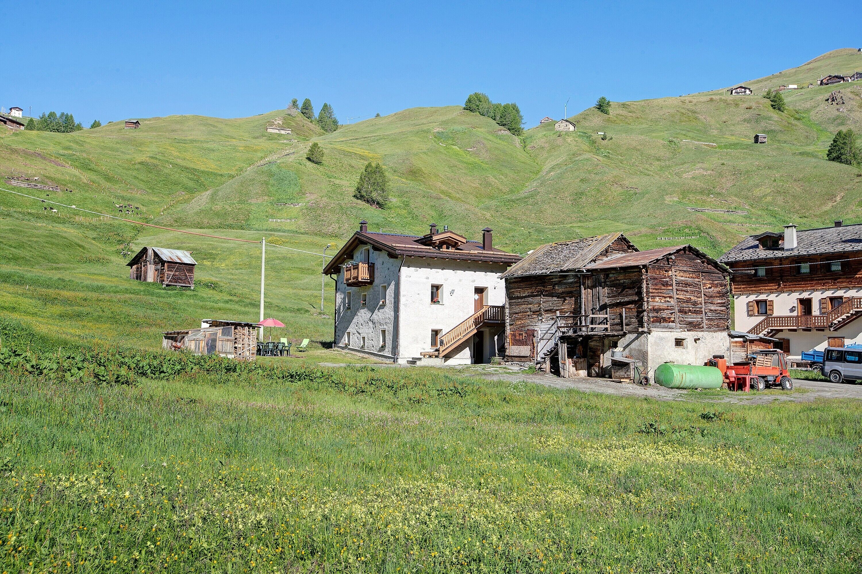 Un village rural avec des maisons traditionnelles en bois, un petit bâtiment doté d’un escalier et un objet cylindrique vert posé sur le sol.
