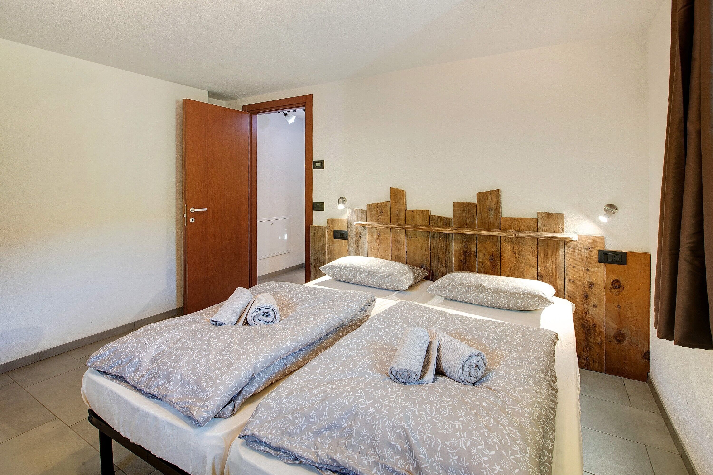 Hotellihuone, jossa on kaksi sänkyä, puinen pääty ja ovi sekä valokytkin seinässä.