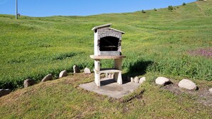 A stone-built outdoor fireplace with a bench, surrounded by rocks and grass on a hill.