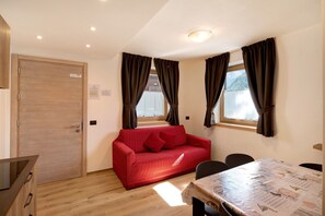 Une pièce avec une porte en bois, un canapé rouge, une table à manger avec des chaises et une fenêtre avec des rideaux.
