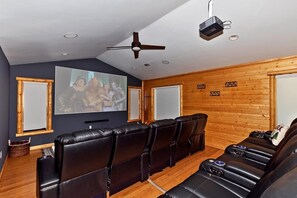 A home theater room with a large screen, leather reclining chairs, wooden walls, and a ceiling fan. - Mountain Chic by Destination Big Bear : Gorgeous Log Home Located in Castle Glen Estates! Game Room! Lake Views! (Big Bear Lake)