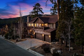 A two-story log cabin with a stone foundation, a garage, and a front porch. - Mountain Chic by Destination Big Bear : Gorgeous Log Home Located in Castle Glen Estates! Game Room! Lake Views! (Big Bear Lake)