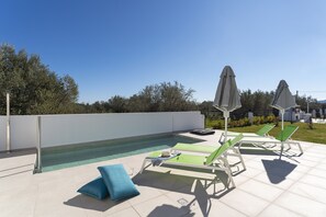 An outdoor swimming pool area with lounge chairs and umbrellas, surrounded by a low wall and greenery.