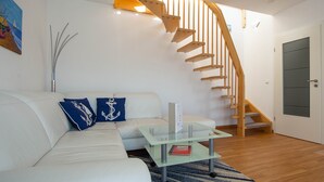 Un salón moderno con un sofá blanco, cojines azules, una mesa de centro de vidrio y una escalera de madera.
