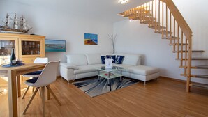Una sala de estar moderna con una escalera de madera, un sofá blanco, una mesa de comedor con sillas y un mueble aparador con puertas de cristal.