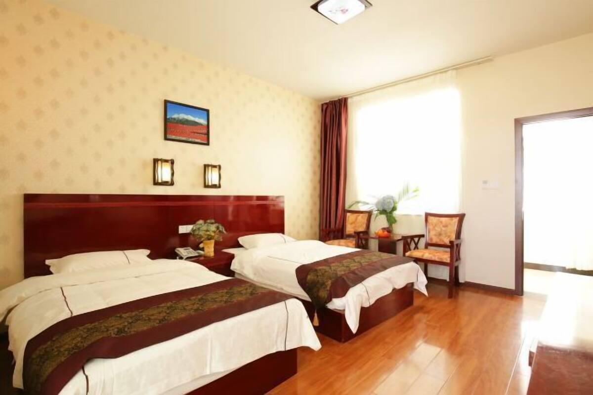 Habitació d'hotel amb dos llits, sòl de fusta, una finestra gran amb cortines, una taula petita amb una cadira i un quadre a la paret.