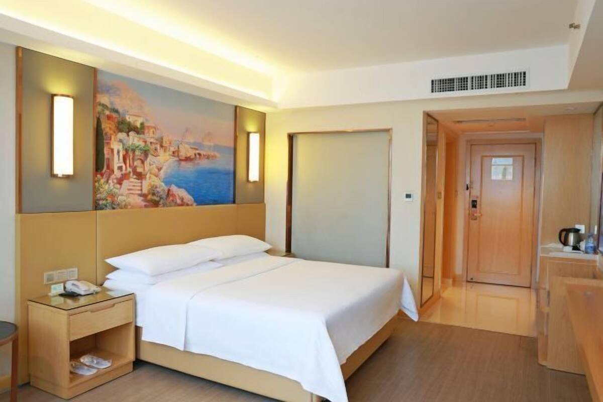 A hotel room with a bed, bedside table, lamp, painting, and a door leading to another room.