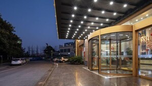 Ein Hotel mit gebogenem Glasfassade, einem überdachten Außenbereich mit Deckenleuchten und einem Parkplatz mit Autos.