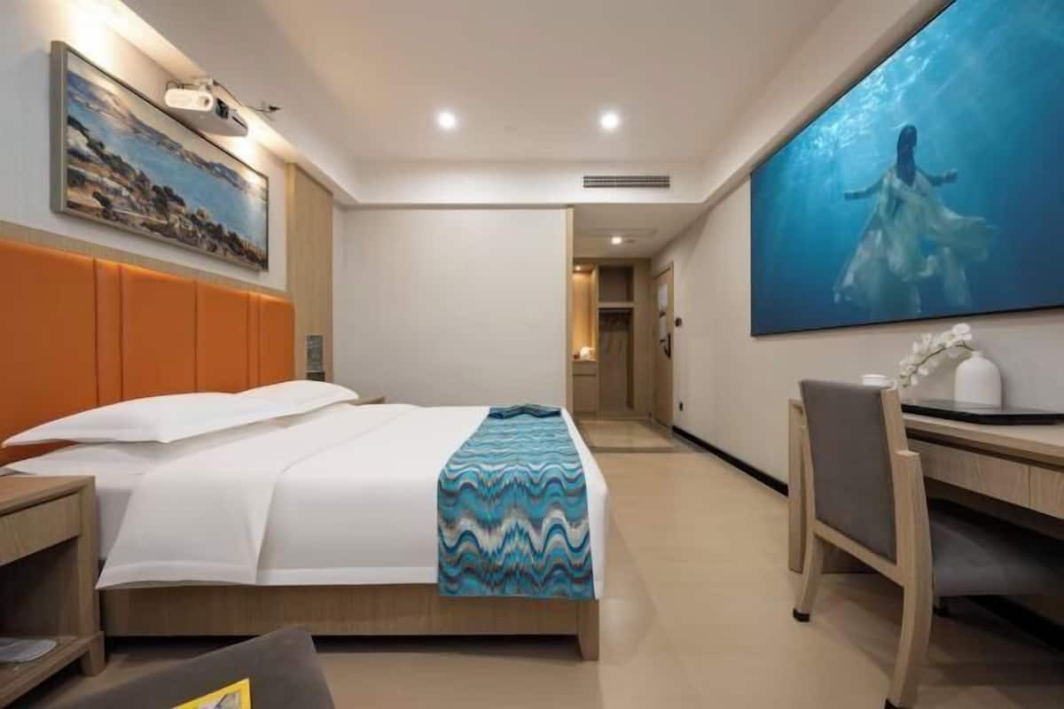 A hotel room with a bed, a desk, and a chair. There is a painting on the wall and a window with a view.