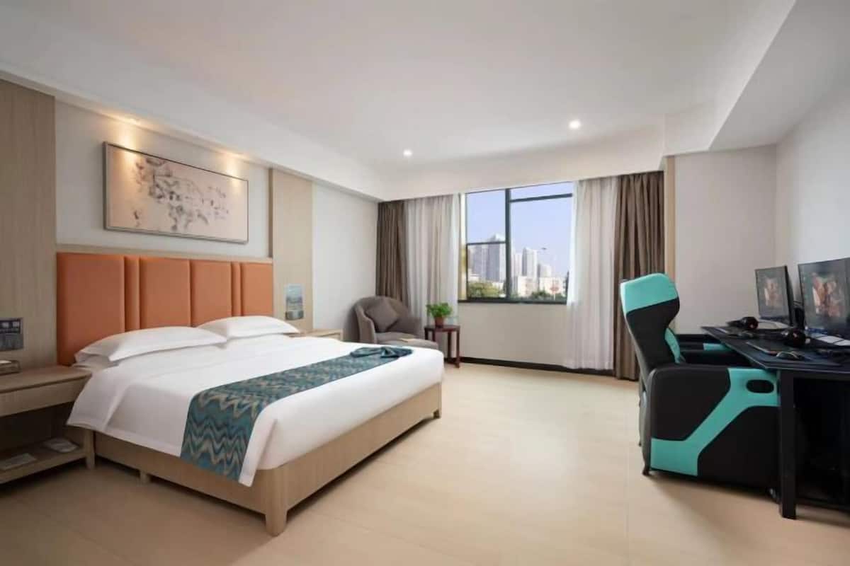 A hotel room with a bed, a desk with a computer, a chair, a window with a city view, and a painting on the wall.