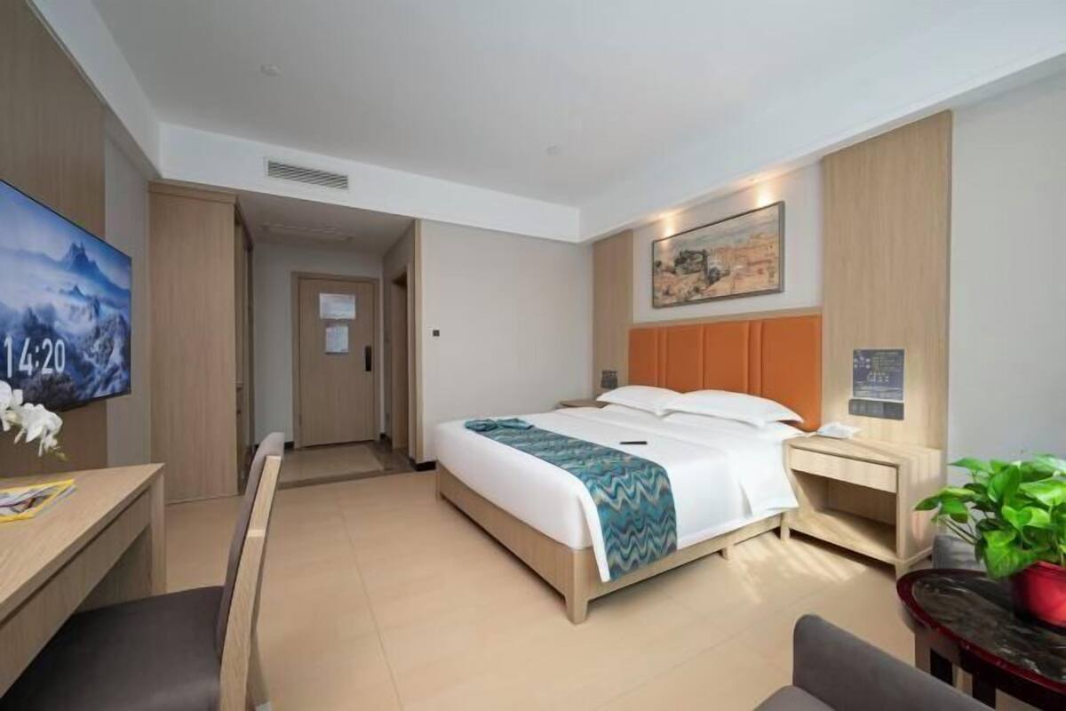 A hotel room with a bed, bedside table, a desk with a chair, a TV, and a plant.