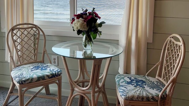 Una stanza con vista sul mare, due poltrone in vimini con cuscini decorati, un tavolino in vetro con un vaso di fiori e tende color beige.