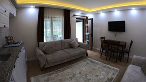 A living room with a sofa, a dining table with chairs, a TV mounted on the wall, and a kitchen area with a gas stove. - Uzungol Adiloglu Apart (Çaykara)