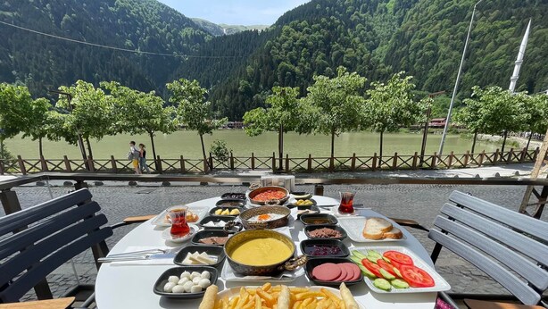 A table set with a variety of dishes, including French fries, bread, and vegetables, overlooking a scenic lake with mountains in the background. - Uzungol Adiloglu Apart (Çaykara)
