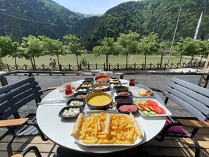 A table set with a variety of dishes, including French fries, bread, and vegetables, overlooking a scenic lake with mountains in the background. - Uzungol Adiloglu Apart (Çaykara)