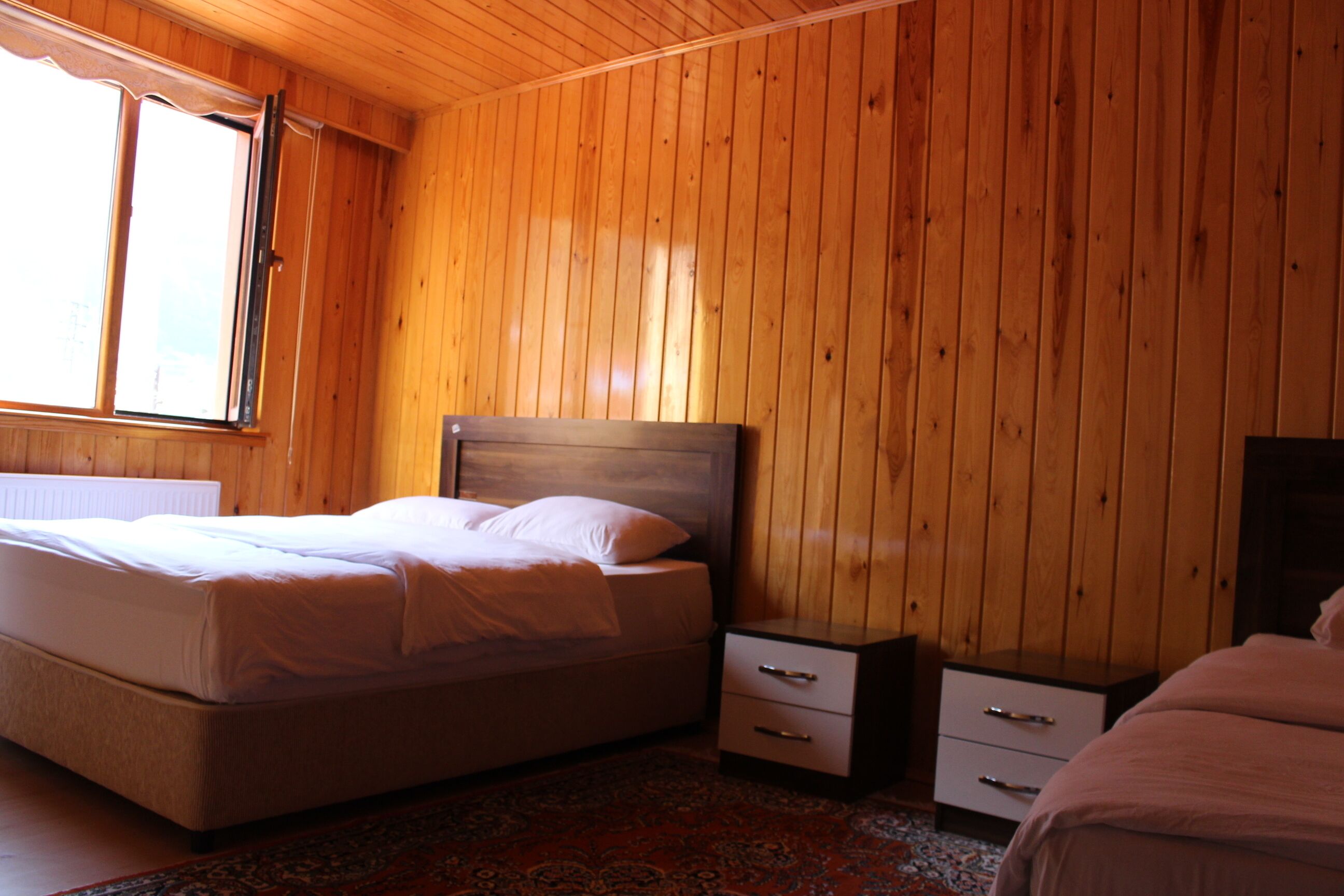 Un dormitorio con pared de paneles de madera, una cama con ropa blanca, una mesita de noche con cajones y un radiador debajo de la ventana.